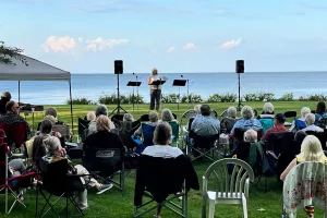 A woman speaks to a crowd of people in chairs outdoors with a lake in the background.